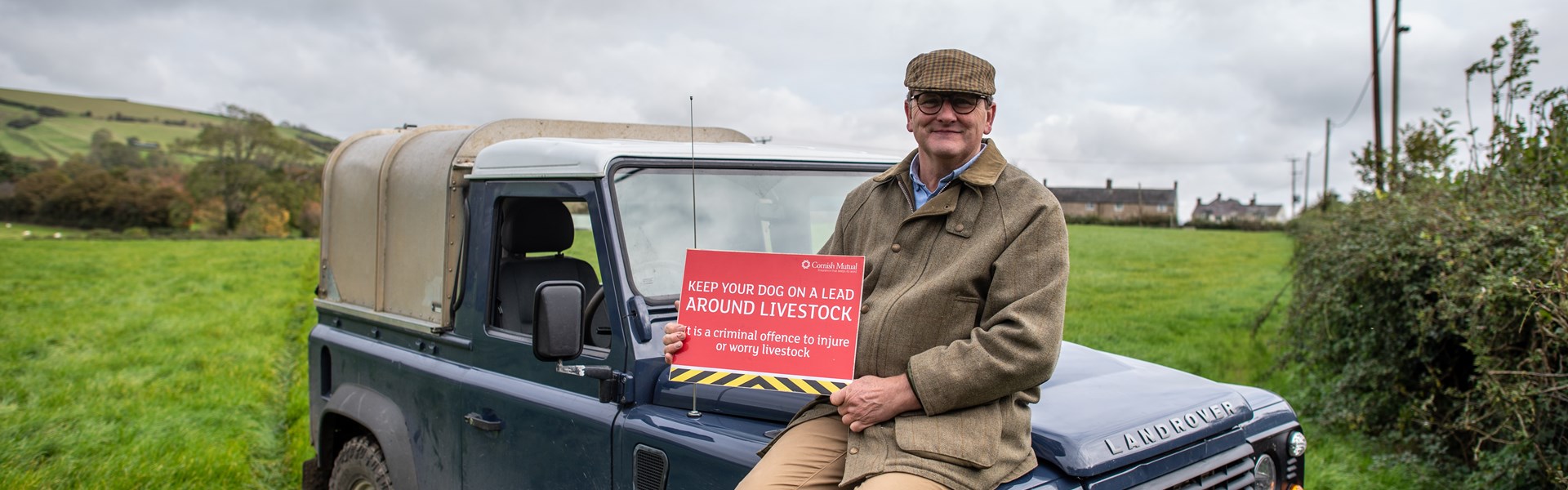 Cornish Mutual Member sat on Land Rover holding Dogs on Lead sign